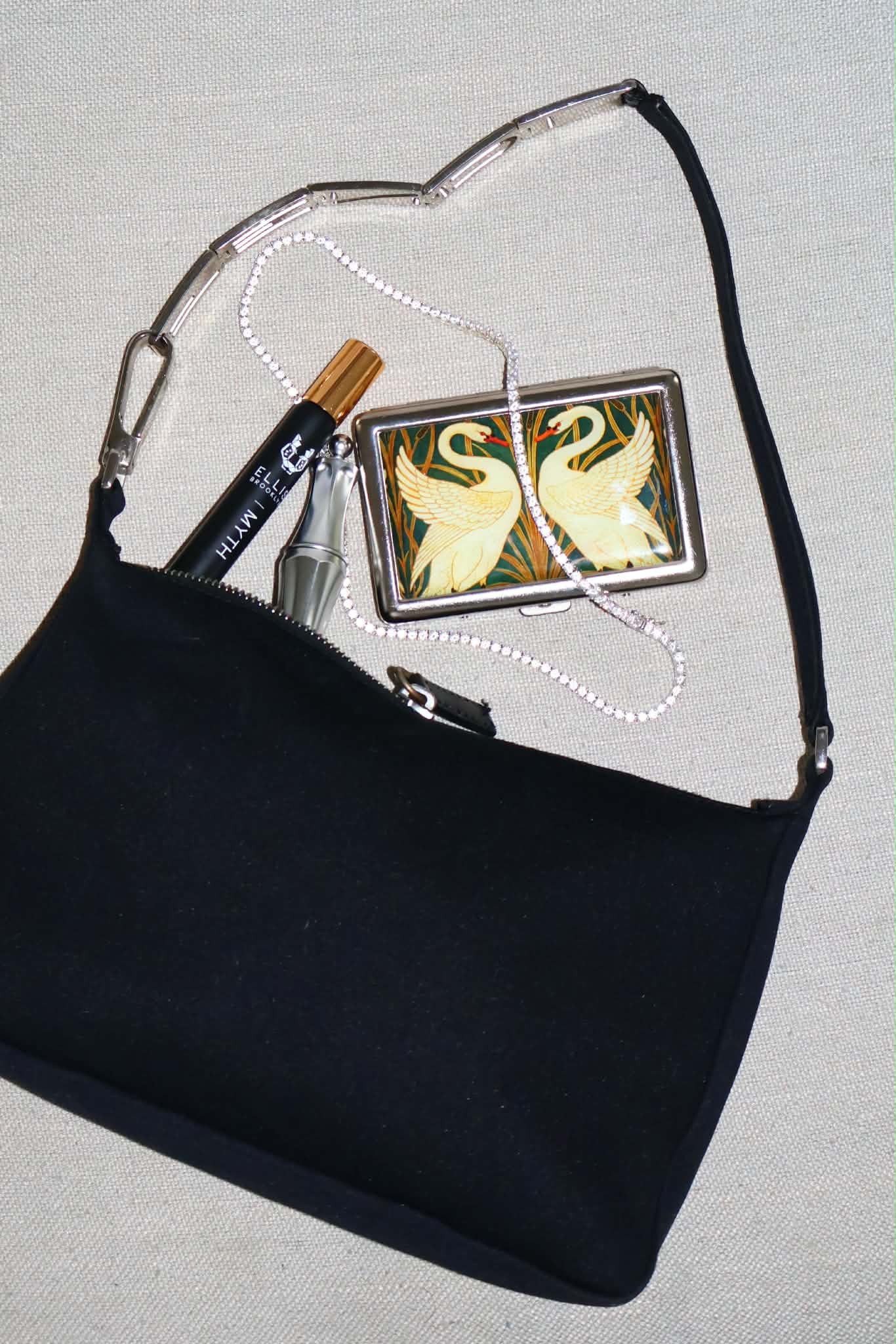 Overhead flatlay of the black shoulder bag on linen with its silver chain strap curled beside a swan cigarette case