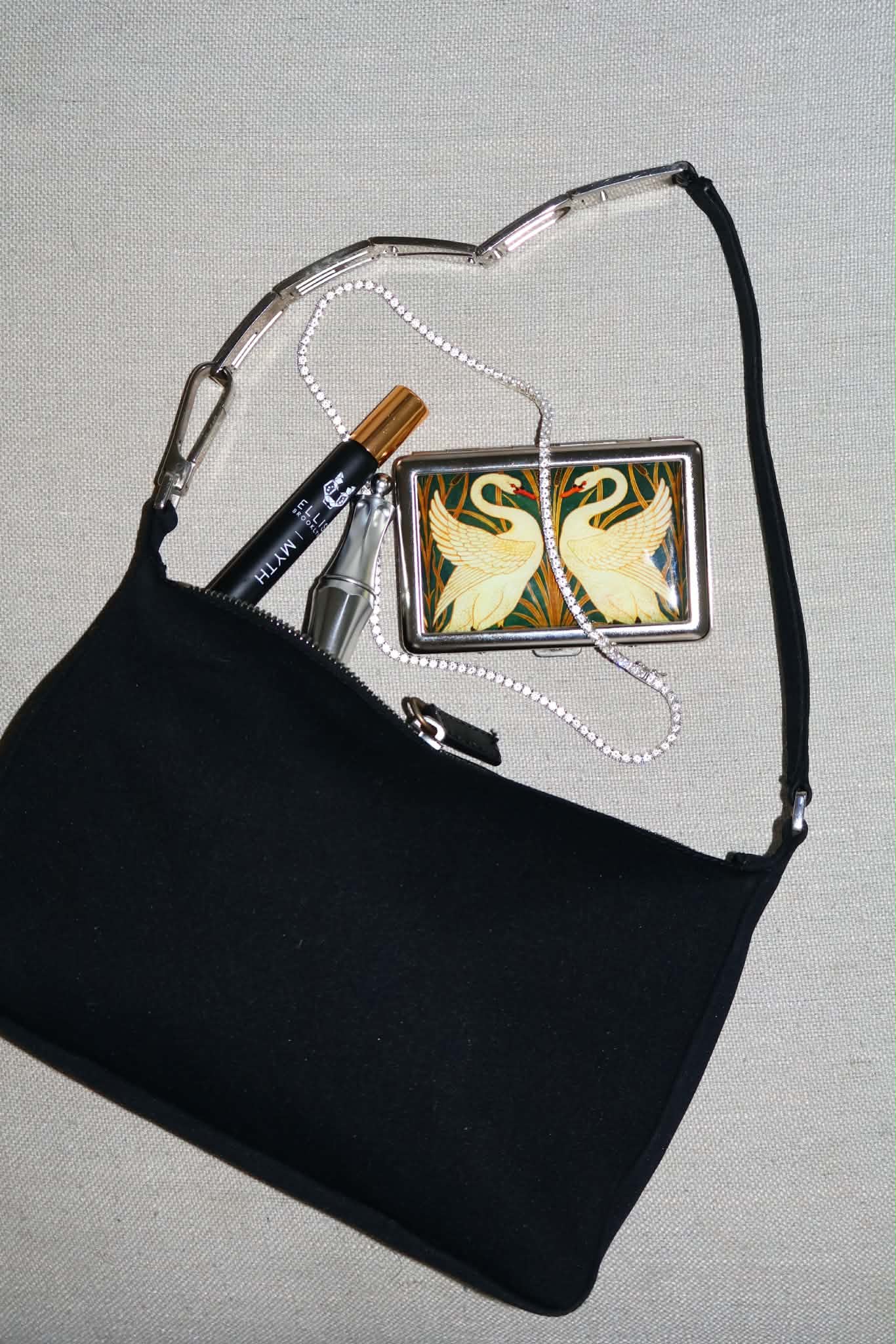 Flatlay of the black bag on linen, unzipped, with perfume and a swan-print case set beside it