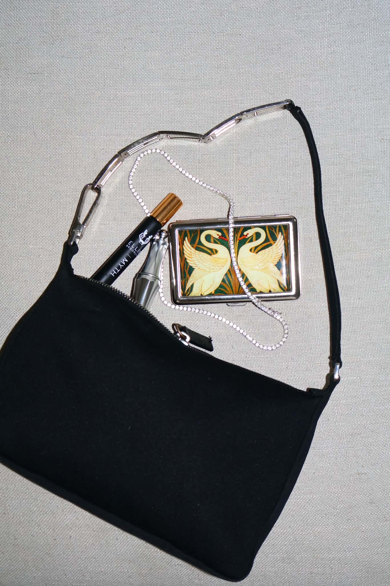 Black satin shoulder bag opened on linen, a swan-print cigarette case, a perfume vial and a rhinestone necklace spilling out
