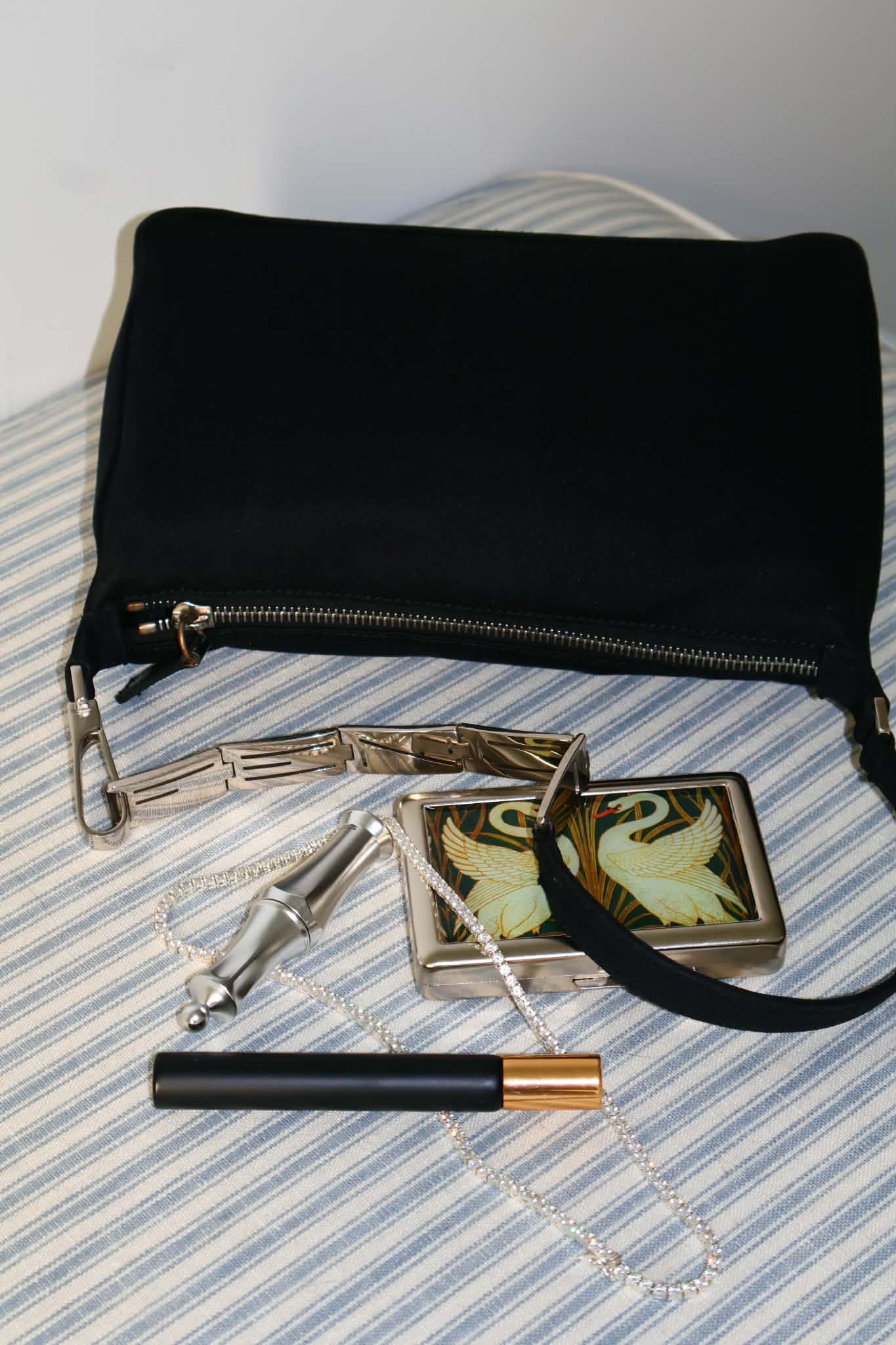The black shoulder bag sitting upright on a striped bedspread, strap and trinkets in front of it