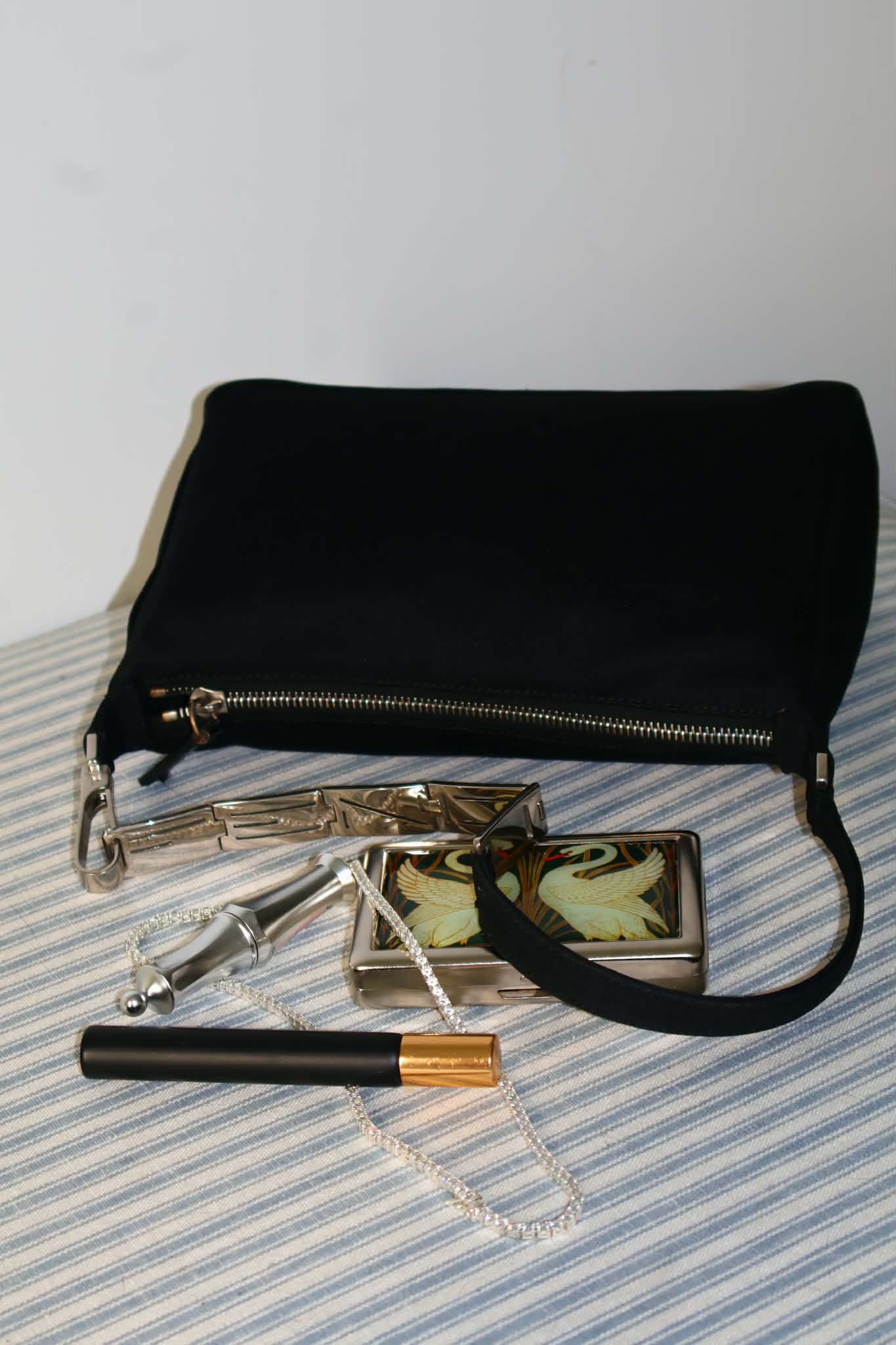 Black satin Fendi shoulder bag with a silver logo-plate strap, photographed on striped ticking beside a swan cigarette case and a perfume vial