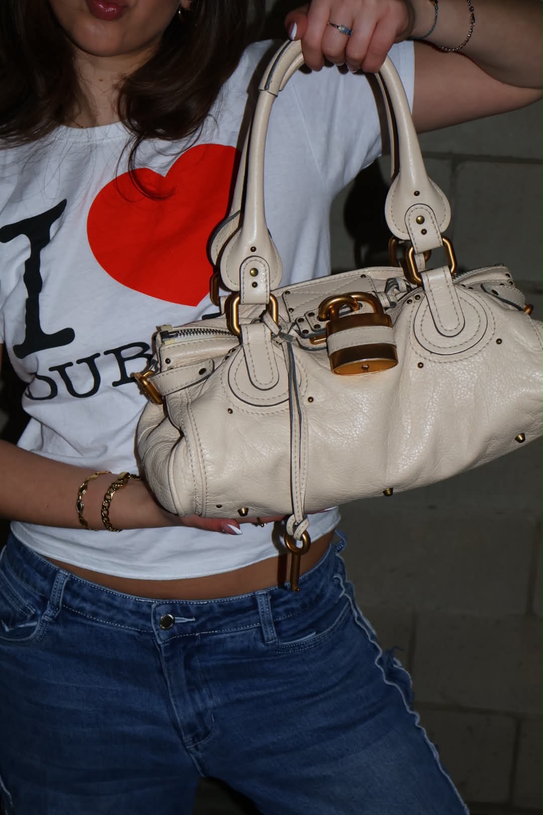The Paddington lifted by its handles across a white tee, gold chain bracelets stacked on the wrist