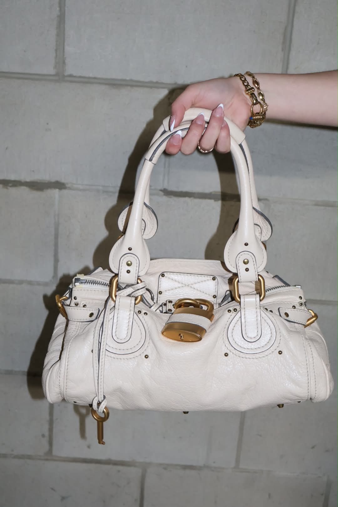 Cream Chloé Paddington bag held by both handles against a painted block wall, showing the brushed gold padlock and studded pebbled leather