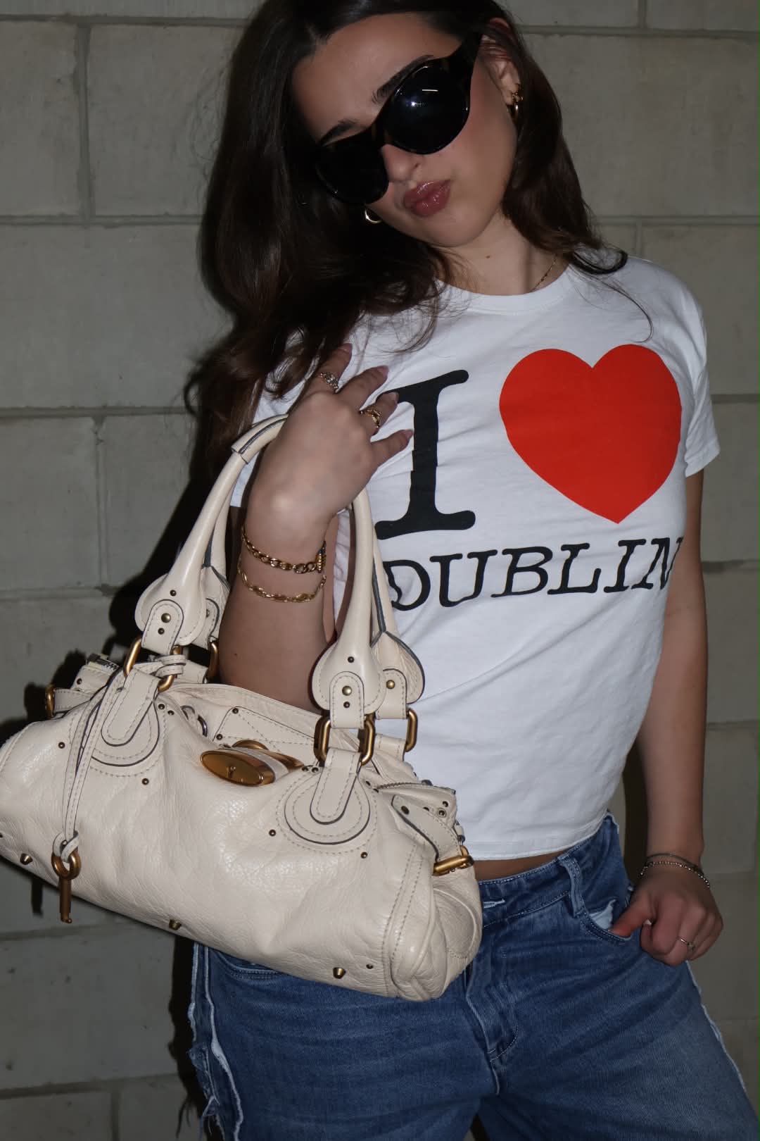 Cream Chloé Paddington held at the hip in dark sunglasses and an I heart Dublin tee, lit hard against a block wall